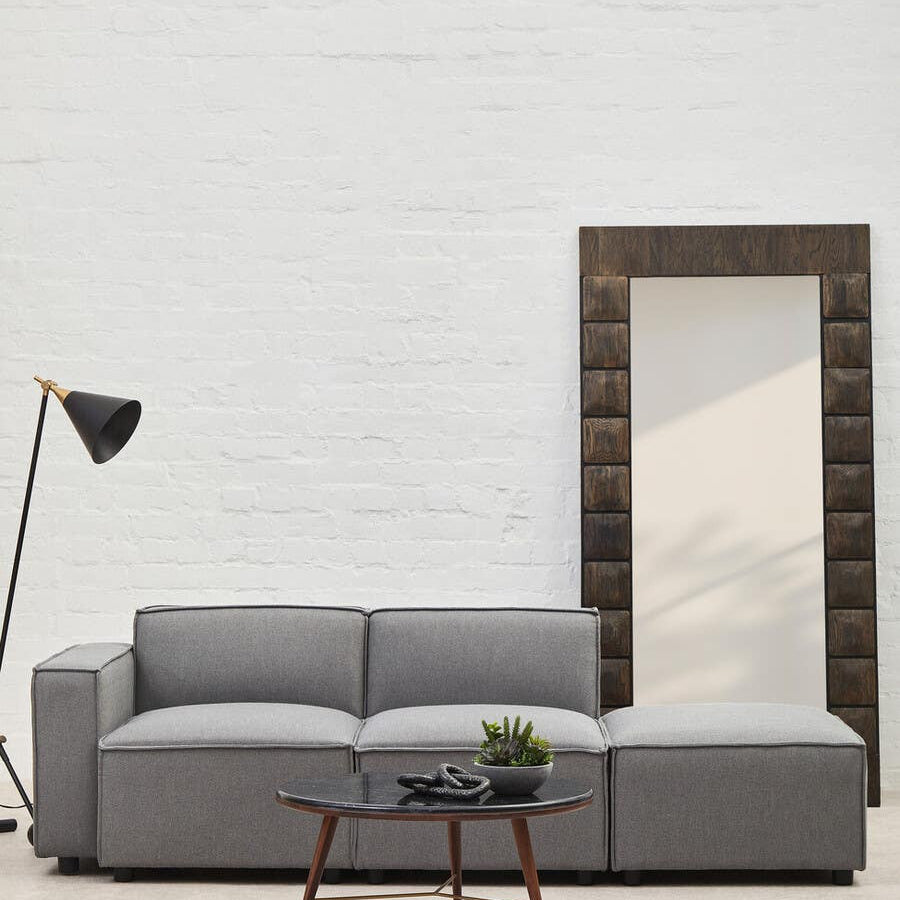 Grey fabric right arm modular sofa with plush cushions, positioned next to a tall mirror and a modern floor lamp, set against a light-colored wall. A wooden coffee table is placed in front of the sofa