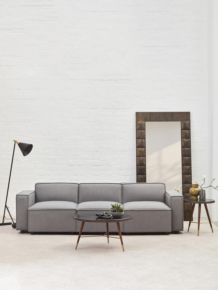 A grey fabric armless modular sofa with three cushions, positioned in a bright room featuring a large mirror and a floor lamp. A small potted plant and a side table are visible next to the sofa, with a light-colored floor and white walls in the background