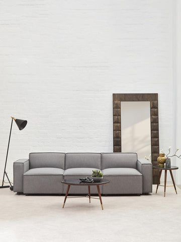 A grey fabric armless modular sofa with three cushions, positioned in a bright room featuring a large mirror and a floor lamp. A small potted plant and a side table are visible next to the sofa, with a light-colored floor and white walls in the background