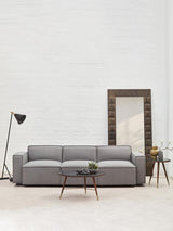 A grey fabric armless modular sofa with three cushions, positioned in a bright room featuring a large mirror and a floor lamp. A small potted plant and a side table are visible next to the sofa, with a light-colored floor and white walls in the background