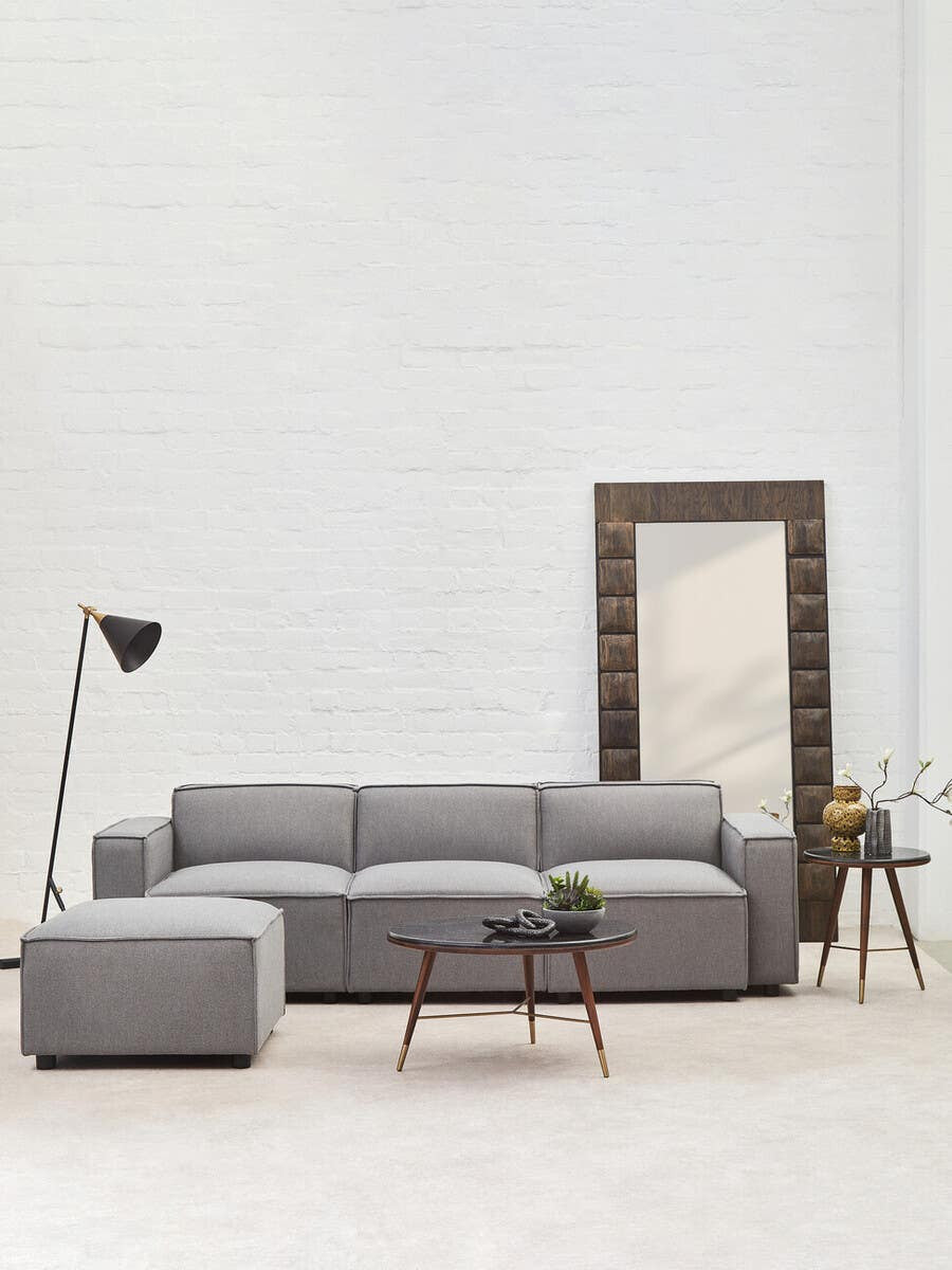 A grey fabric armless modular sofa is positioned against a light-colored wall, accompanied by a tall mirror with a decorative frame, a small round side table, and a floor lamp with a black shade. A small potted plant sits on the side table, and the floor is covered with a light-colored rug