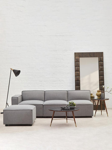 Grey fabric armless modular sofa positioned in a bright room with a large mirror and a wooden coffee table. A floor lamp stands beside the sofa, and a decorative plant is placed on the table. The walls are light-colored, enhancing the modern aesthetic of the space