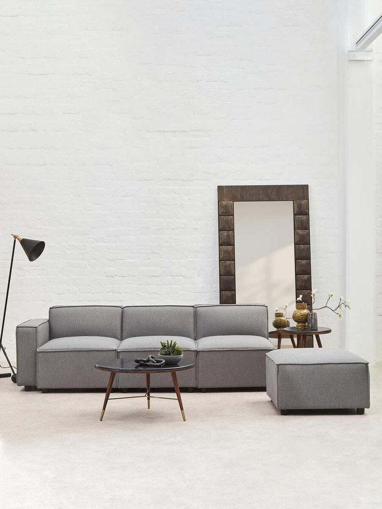 Grey fabric armless modular sofa positioned in a bright, minimalist living space, accompanied by a small square ottoman, a wooden coffee table with decorative items, a floor lamp, and a large mirror reflecting the room's light