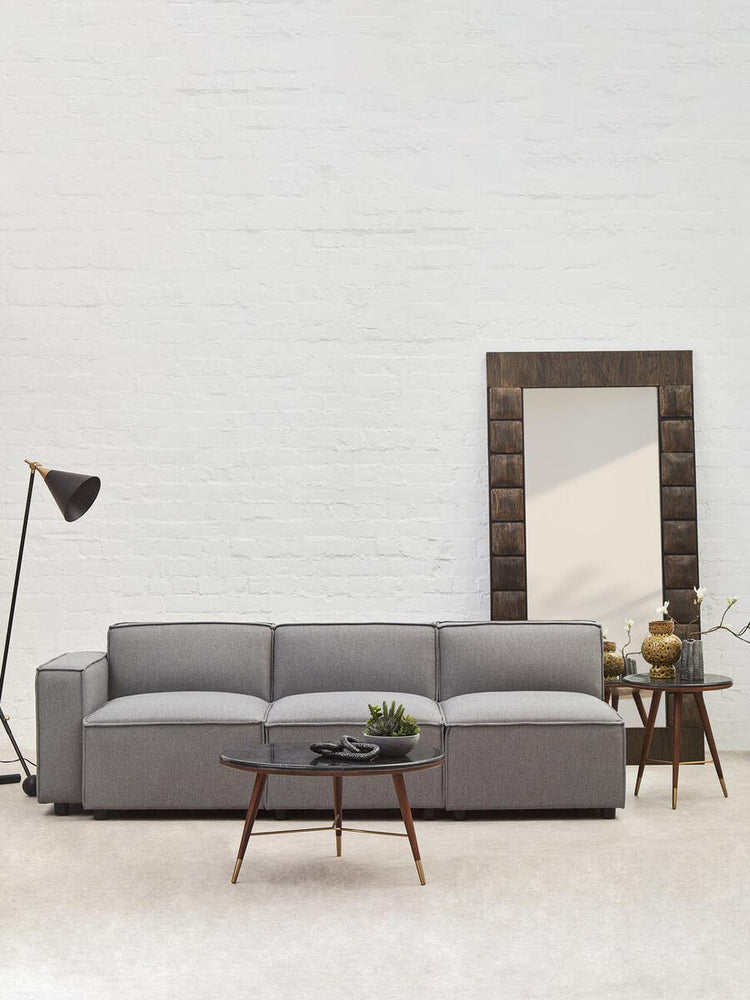 Grey fabric armless modular sofa with three plush cushions, set against a light-colored wall. A tall floor lamp stands to the left, and a decorative mirror with a wooden frame is positioned behind the sofa. A small potted plant is placed on a side table next to the sofa