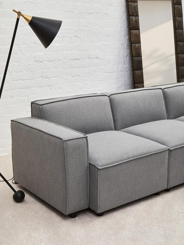 Grey fabric armless modular sofa with a sleek design, featuring three cushioned sections and a minimalist aesthetic. A black floor lamp stands nearby, and a large mirror is visible in the background against a light-colored wall. The sofa is positioned on a light-colored floor