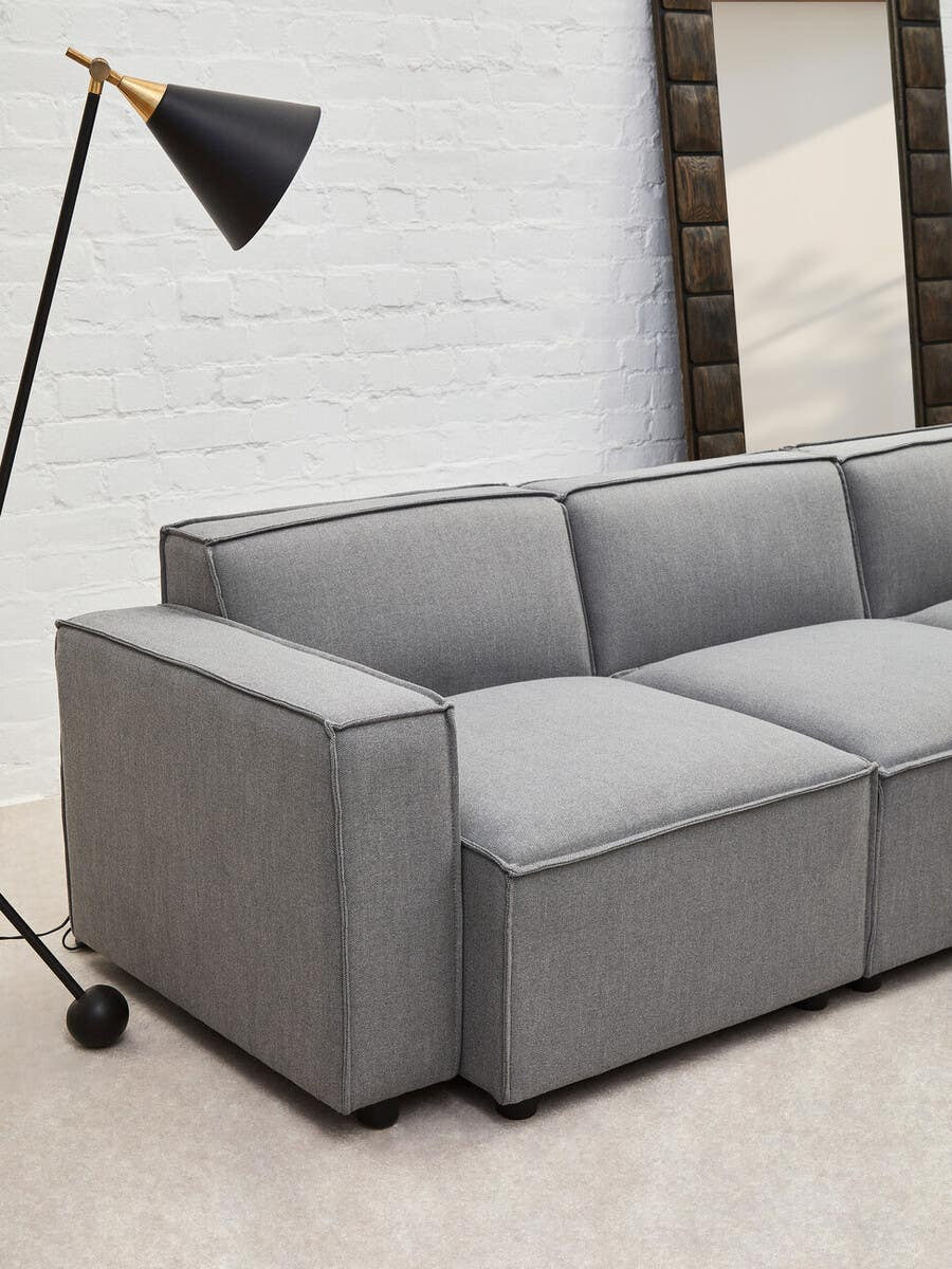 Grey fabric armless modular sofa with a sleek design, featuring three cushioned sections and a minimalist aesthetic. A black floor lamp stands nearby, and a large mirror is visible in the background against a light-colored wall. The sofa is positioned on a light-colored floor