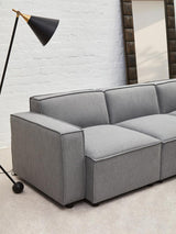 Grey fabric armless modular sofa with a sleek design, featuring three cushioned sections and a minimalist aesthetic. A black floor lamp stands nearby, and a large mirror is visible in the background against a light-colored wall. The sofa is positioned on a light-colored floor
