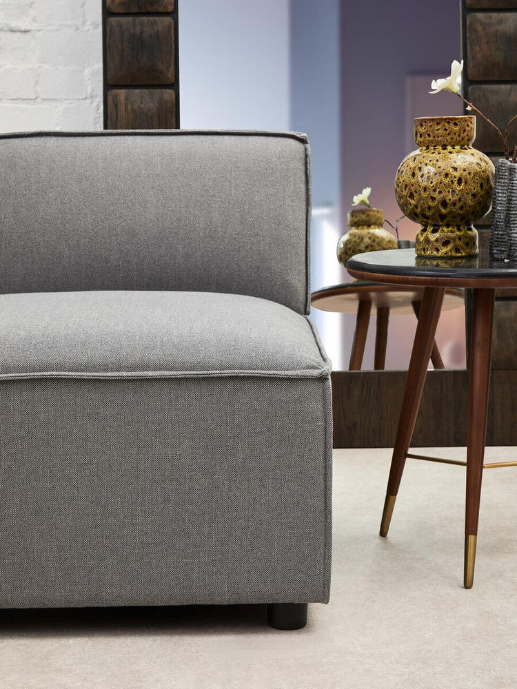 Armless modular sofa in grey fabric with a sleek design, featuring a cushioned seat and a low backrest, positioned next to a wooden side table with a decorative gold vase. The background includes a softly lit room with neutral tones