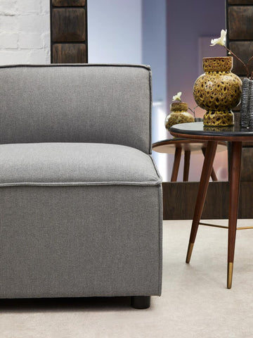 Armless modular sofa in grey fabric with a sleek design, featuring a cushioned seat and a low backrest, positioned next to a wooden side table with a decorative gold vase. The background includes a softly lit room with neutral tones
