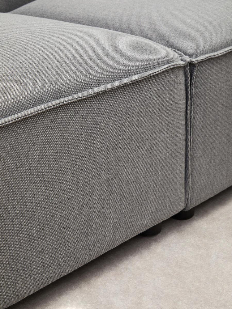 Close-up view of the Toulouse Grey Fabric Armless Modular Sofa, showcasing the soft grey upholstery and clean lines of the modular design, with a focus on the seam and corner details. The surface appears smooth and inviting, set against a light-colored floor
