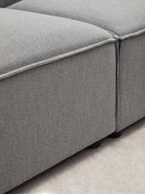 Close-up view of the Toulouse Grey Fabric Armless Modular Sofa, showcasing the soft grey upholstery and clean lines of the modular design, with a focus on the seam and corner details. The surface appears smooth and inviting, set against a light-colored floor
