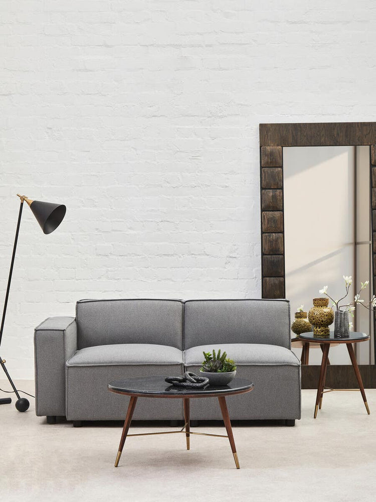A grey fabric armless modular sofa is positioned in a modern living space, accompanied by a wooden side table with decorative items and a floor lamp. A large mirror reflects the room's light, enhancing the contemporary aesthetic. The wall behind is textured and neutral, creating a cozy atmosphere