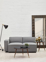 A grey fabric armless modular sofa is positioned in a modern living space, accompanied by a wooden side table with decorative items and a floor lamp. A large mirror reflects the room's light, enhancing the contemporary aesthetic. The wall behind is textured and neutral, creating a cozy atmosphere