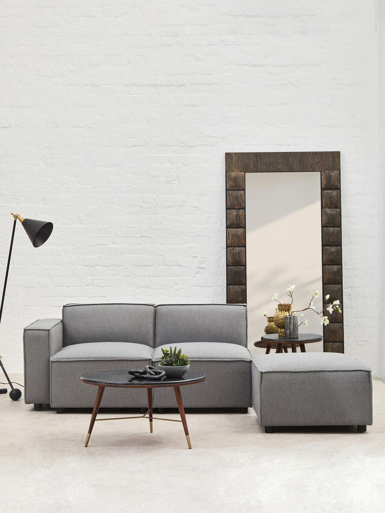 Grey fabric armless modular sofa with a modern design, accompanied by a matching ottoman, set against a white wall. A floor lamp and a wooden coffee table with decorative items are also visible in the scene, along with a large mirror reflecting the room