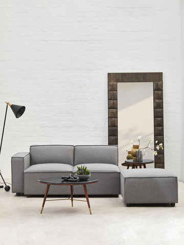 Grey fabric armless modular sofa with a modern design, accompanied by a matching ottoman, set against a white wall. A floor lamp and a wooden coffee table with decorative items are also visible in the scene, along with a large mirror reflecting the room
