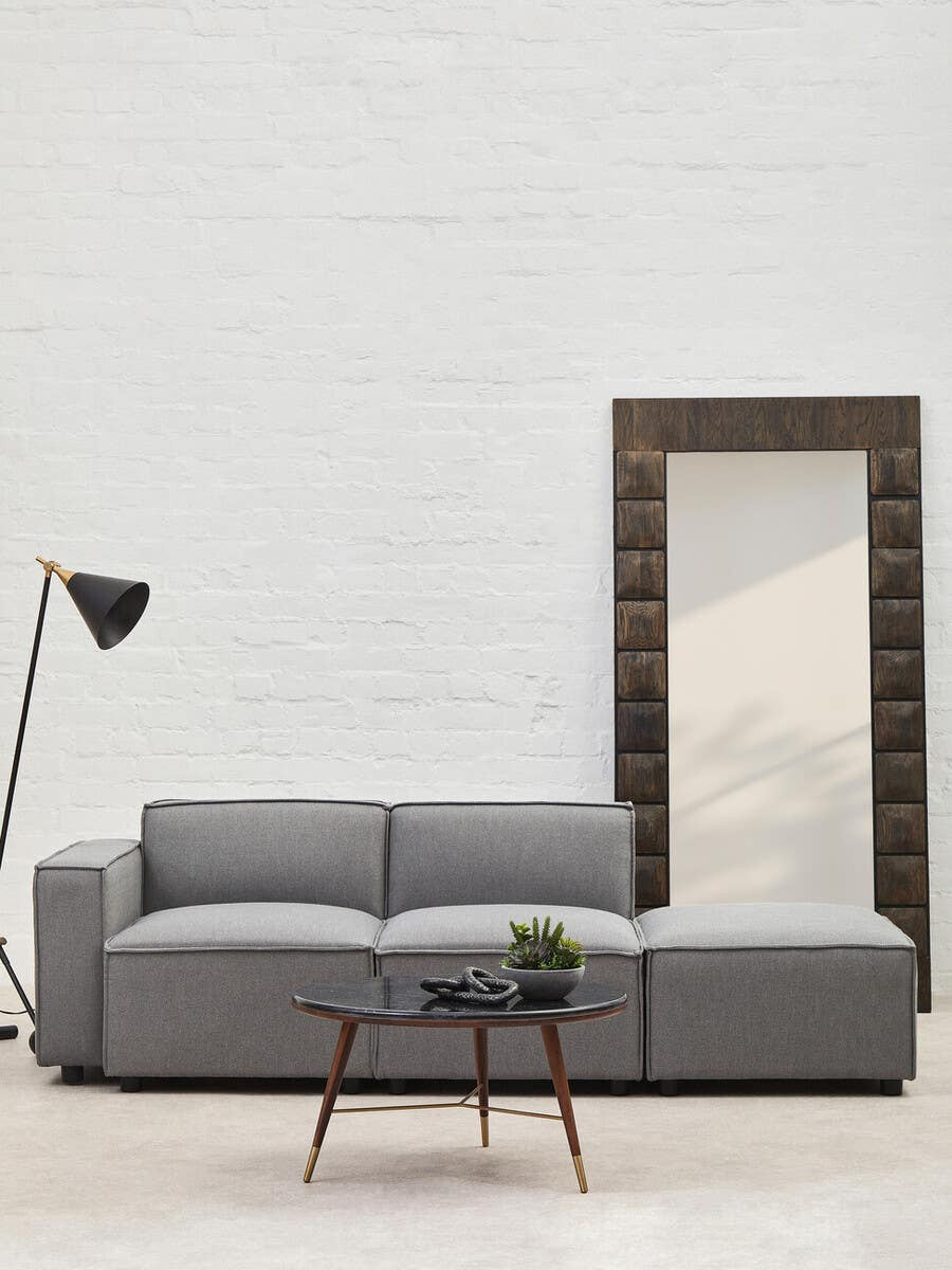 Grey fabric armless modular sofa with plush cushions, positioned next to a tall mirror and a modern floor lamp, set against a light-colored wall and a wooden coffee table