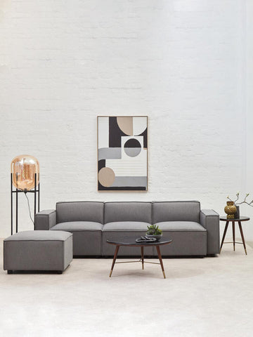Grey fabric armless modular sofa positioned in a modern living space, accompanied by a decorative lamp and a small side table with a plant. A large wall mirror reflects the room's light, enhancing the contemporary aesthetic. The sofa features clean lines and plush cushions, creating a cozy yet stylish atmosphere
