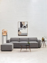 Grey fabric armless modular sofa positioned in a modern living space, accompanied by a decorative lamp and a small side table with a plant. A large wall mirror reflects the room's light, enhancing the contemporary aesthetic. The sofa features clean lines and plush cushions, creating a cozy yet stylish atmosphere