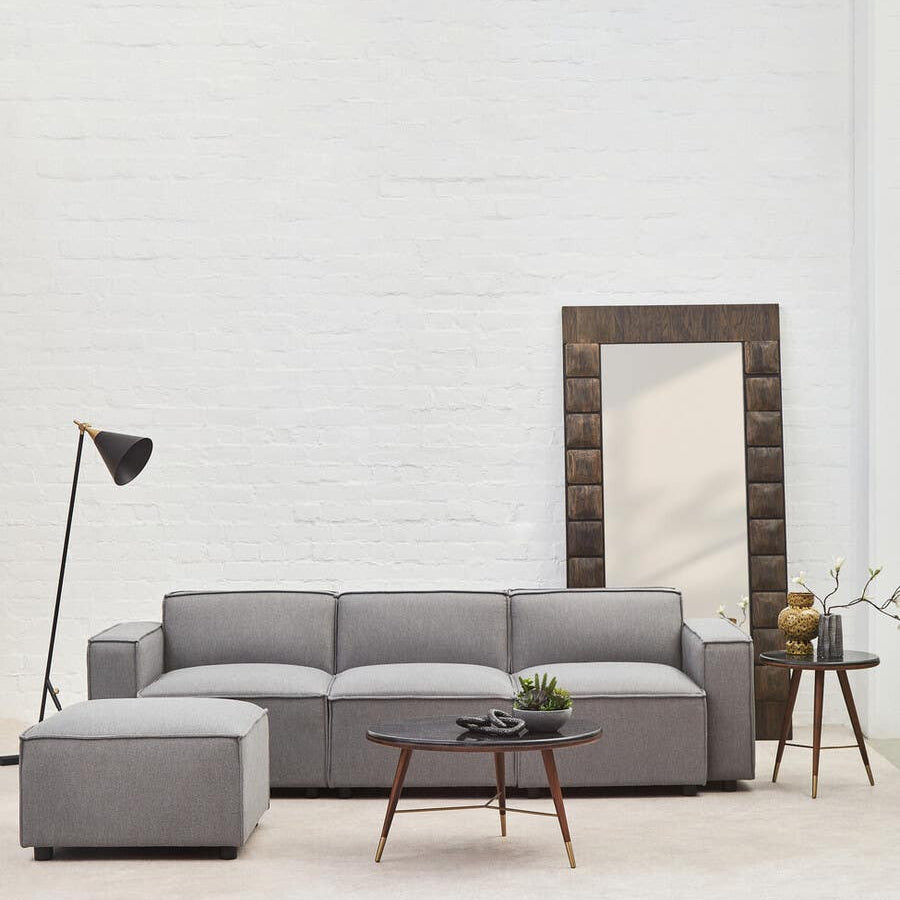 A modern grey fabric left arm modular sofa is positioned in a bright, minimalist living space. The sofa features plush cushions and a sleek design, complemented by a small round coffee table with a plant on it. A tall floor lamp stands nearby, and a large mirror with a decorative frame leans against the wall, enhancing the room's aesthetic. The floor is light-colored, contributing to the airy atmosphere