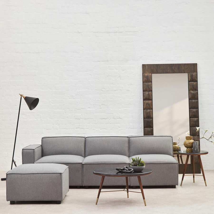 Grey fabric left arm modular sofa with a contemporary design, featuring plush cushions and a sleek silhouette. Accompanied by a small side table and a floor lamp, set against a light-colored wall and a large decorative mirror