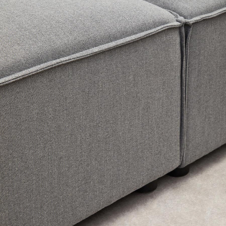 Close-up view of the corner of a grey fabric modular sofa, showcasing the textured upholstery and clean lines of the design, with a visible seam and a low-profile base. The background features a light-colored floor, enhancing the sofa's modern aesthetic