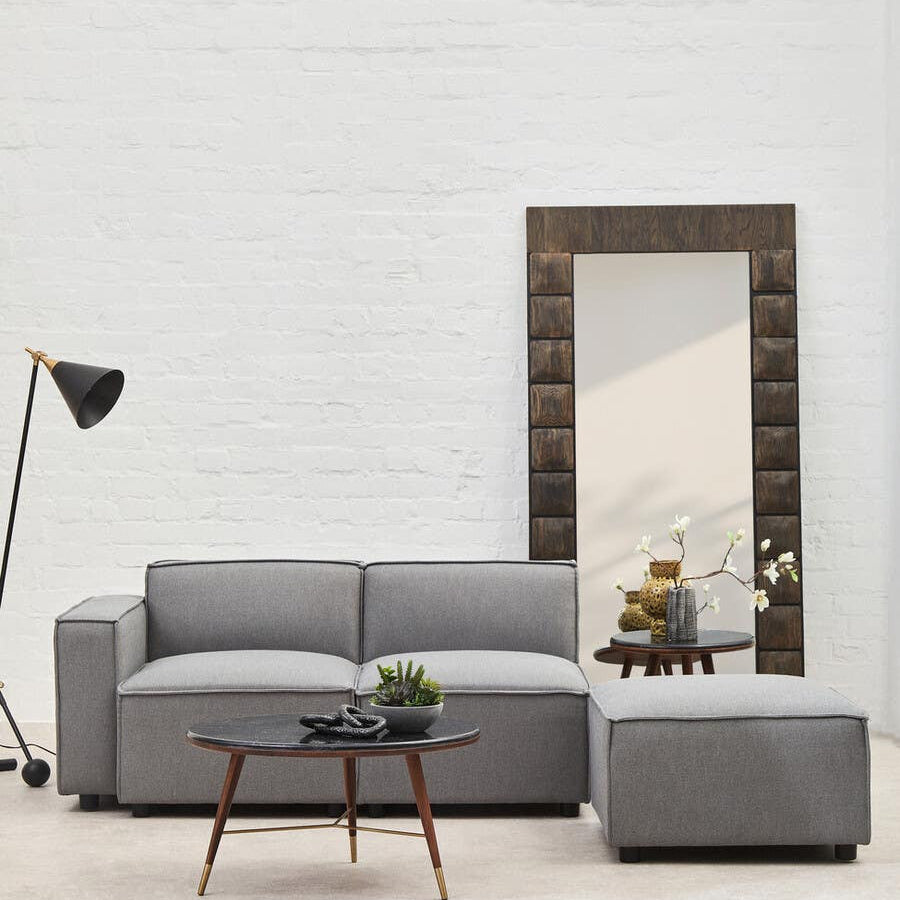 Grey fabric left arm modular sofa with a contemporary design, featuring a spacious seating area and clean lines. Accompanied by a matching ottoman, a wooden coffee table with a decorative plant, and a tall floor lamp. A large mirror is positioned against the wall, reflecting the room's light and space. The background consists of a plain white wall, enhancing the modern aesthetic