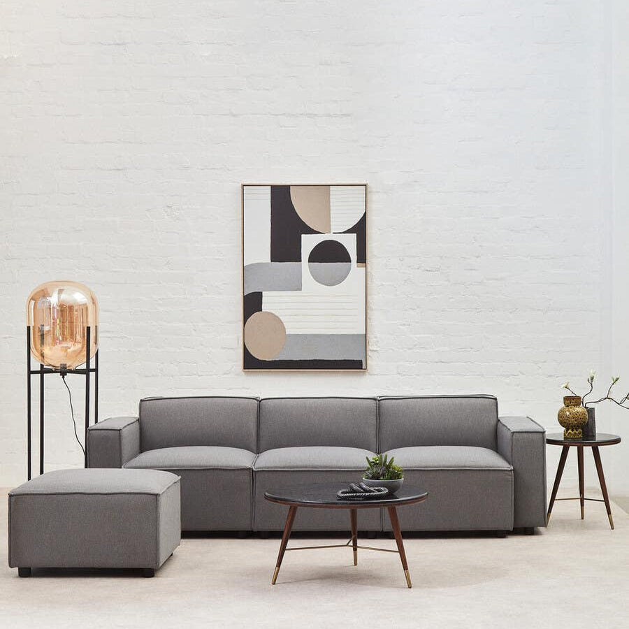 Grey fabric left arm modular sofa with plush cushions, positioned in a modern living space. A large wall mirror hangs above the sofa, reflecting light and enhancing the room's brightness. A small side table with a decorative lamp and a potted plant are placed nearby, adding to the contemporary decor. The background features a light-colored wall, creating a clean and airy atmosphere