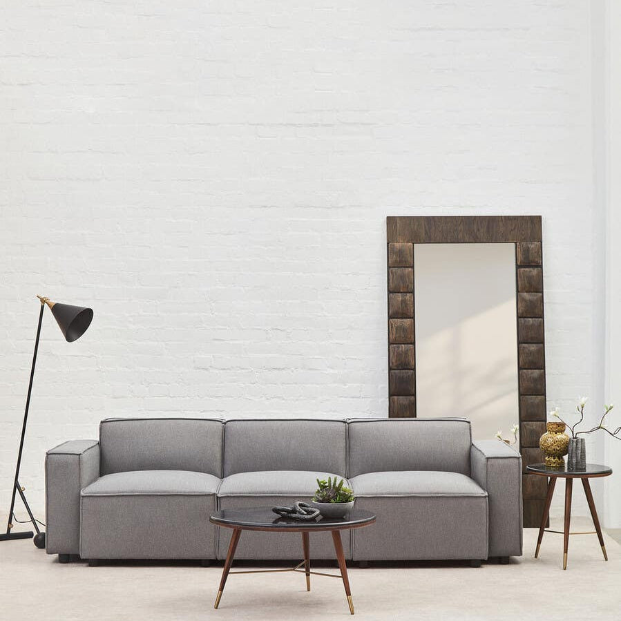 Grey fabric left arm modular sofa with three cushions, positioned in a bright room featuring a large mirror and a small side table with a plant. A floor lamp stands beside the sofa, and the walls are painted white