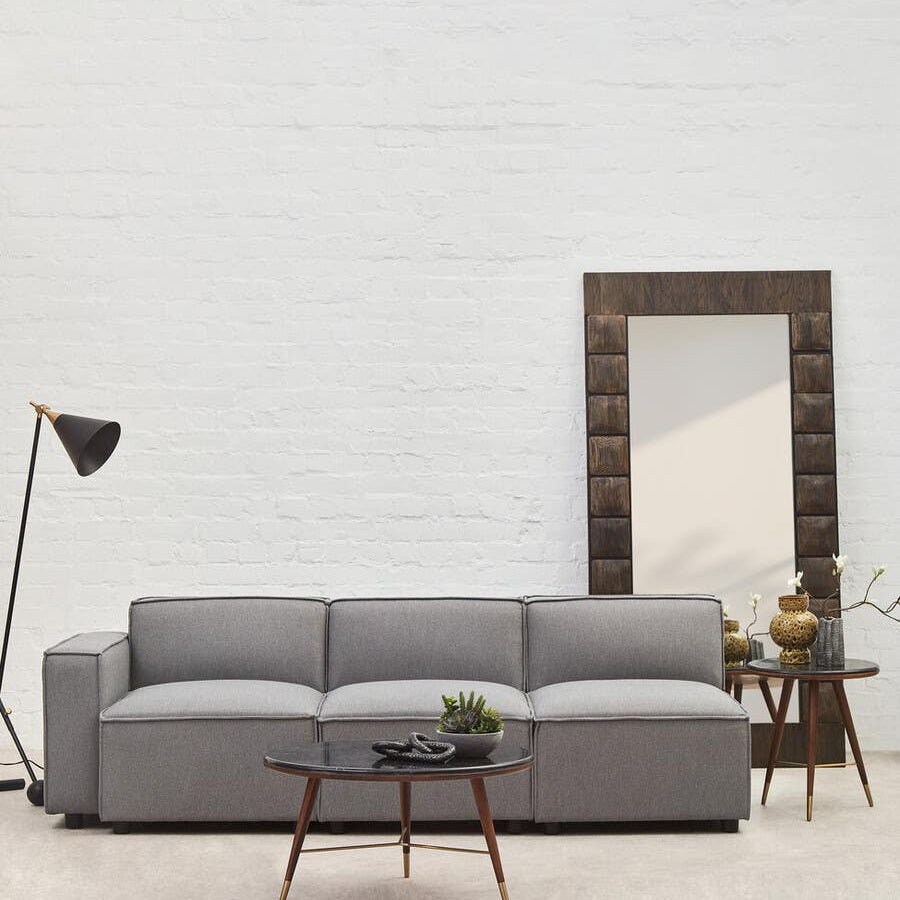 A grey fabric left arm modular sofa with three cushioned seats, resting on wooden legs, positioned in a bright room featuring a large mirror and a floor lamp. A small potted plant is placed on a side table next to the sofa