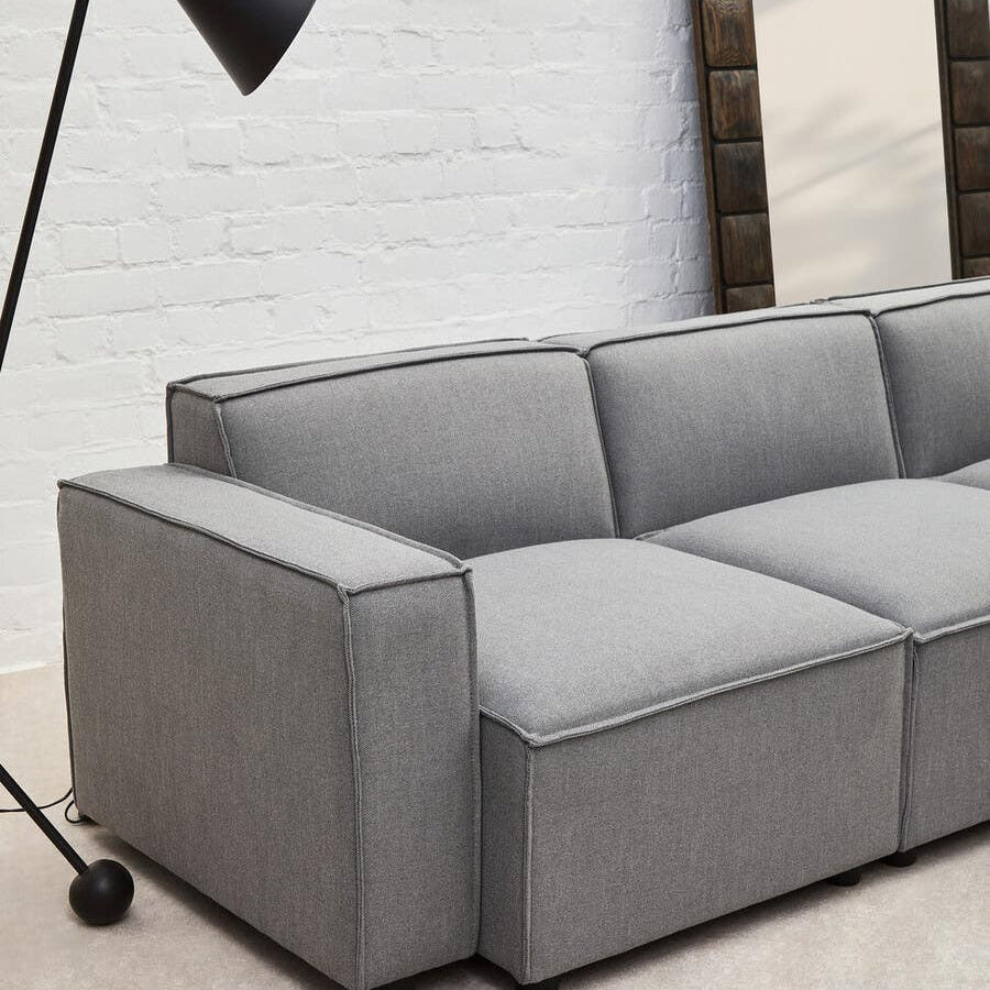 Grey fabric left arm modular sofa with clean lines and a modern design, featuring plush cushions and a sturdy base. A sleek black floor lamp stands nearby, illuminating the space, while a large mirror is mounted on the wall behind the sofa. The flooring is light-colored, enhancing the contemporary aesthetic