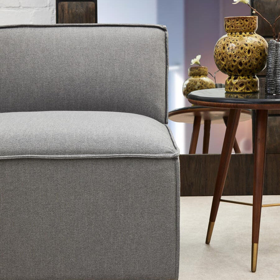 Left arm of a grey fabric modular sofa with a smooth, cushioned seat and a clean, modern design. The sofa is positioned next to a mid-century style wooden side table with a decorative gold vase on top. The background features a softly lit room with neutral-colored walls and a stylish decor