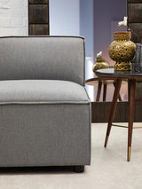 Left arm of a grey fabric modular sofa with a smooth, cushioned seat and a clean, modern design. The sofa is positioned next to a mid-century style wooden side table with a decorative gold vase on top. The background features a softly lit room with neutral-colored walls and a stylish decor