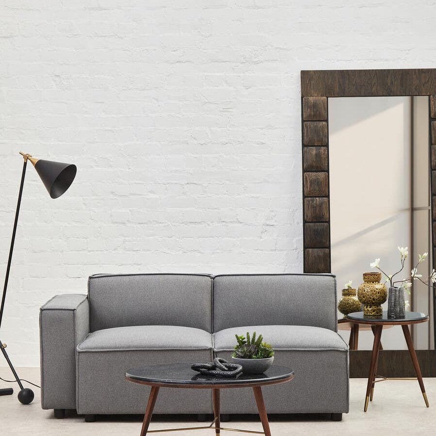 Grey fabric left arm modular sofa with two seat cushions, positioned next to a wooden coffee table and a floor lamp. A large mirror is visible in the background, reflecting the room's light-colored walls and flooring. A small potted plant sits on the coffee table, adding a touch of greenery to the scene