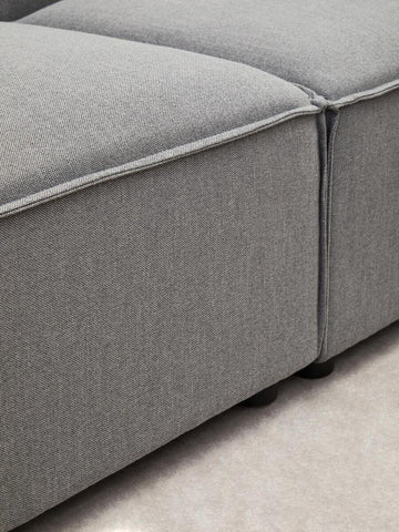 Close-up view of a grey fabric modular ottoman, showcasing its textured upholstery and clean lines. The ottoman features a seamless design with a subtle seam detail and rests on small, dark legs against a light-colored floor