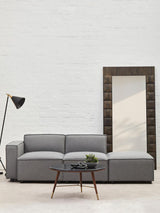 Grey fabric modular ottoman positioned next to a modern sofa, with a floor lamp and a large mirror in the background. The ottoman features a sleek design and is placed on a light-colored floor against a white wall
