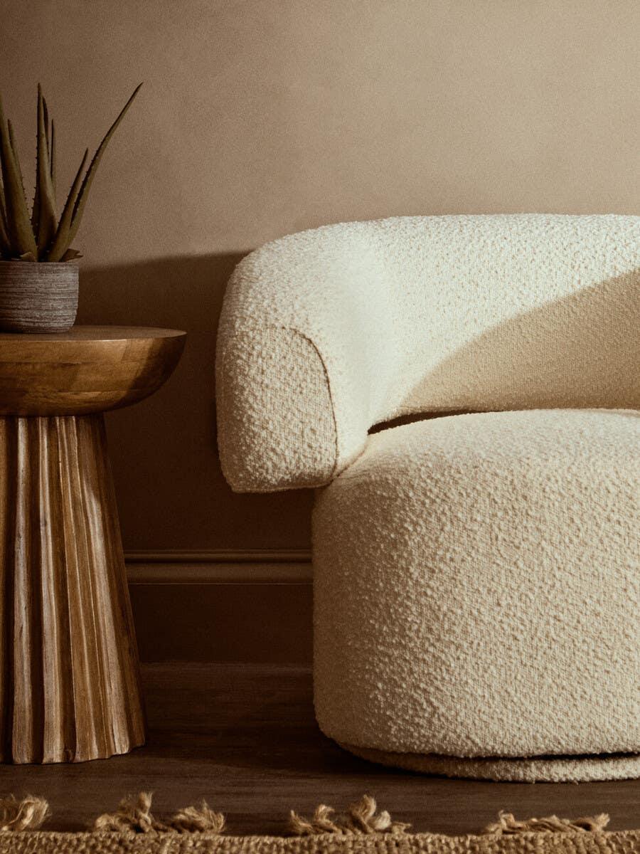 Cream boucle accent chair with curved arm next to wooden side table with plant