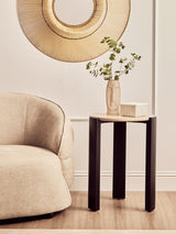 Round side table featuring a cream marble top and sleek black acacia wood legs, positioned next to a soft beige upholstered sofa.