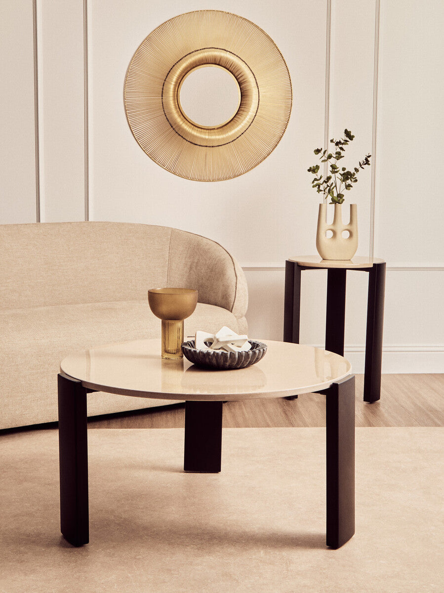 Round side table featuring a cream marble top and sleek black acacia wood legs, set against a soft beige sofa and a decorative gold wall mirror.