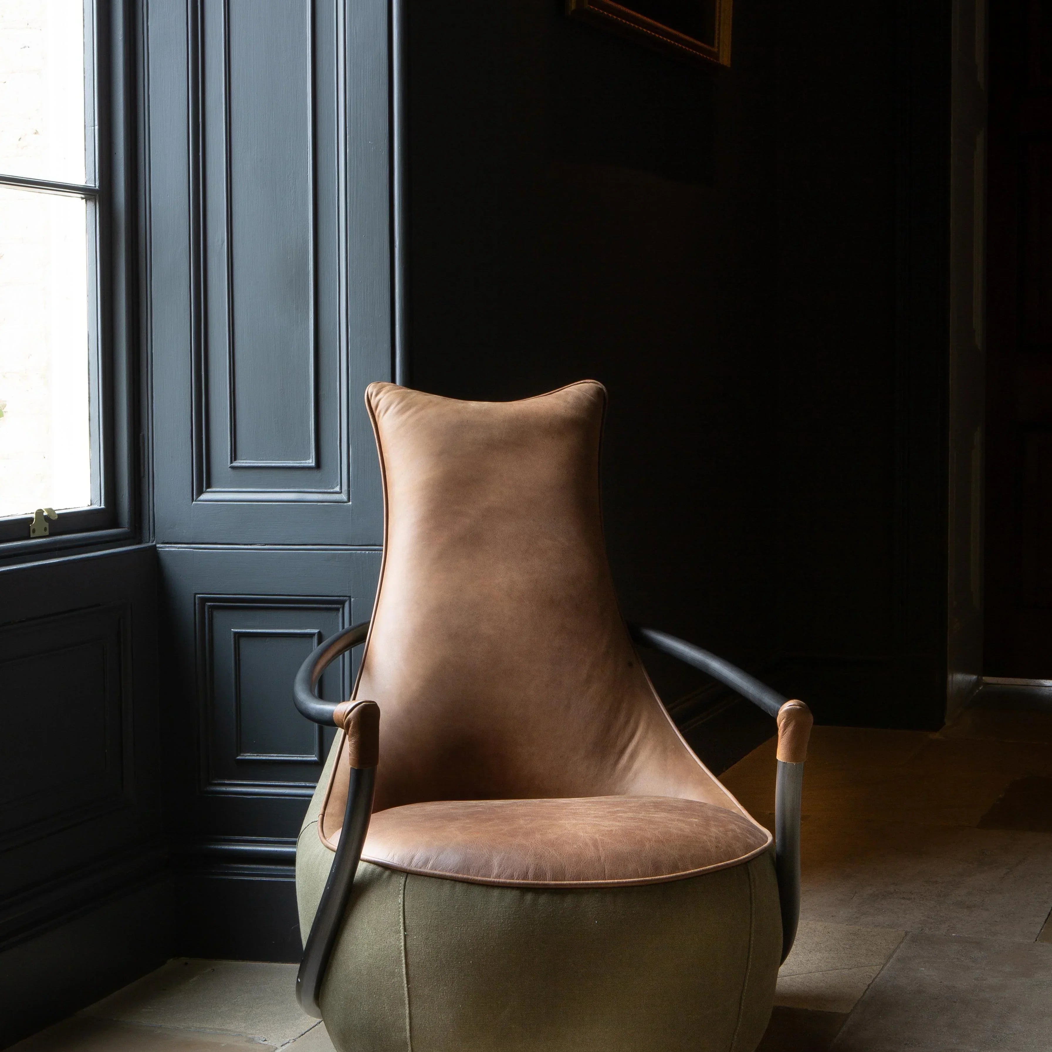 Brown leather accent chair with fabric base and curved arms in a dark room by a window