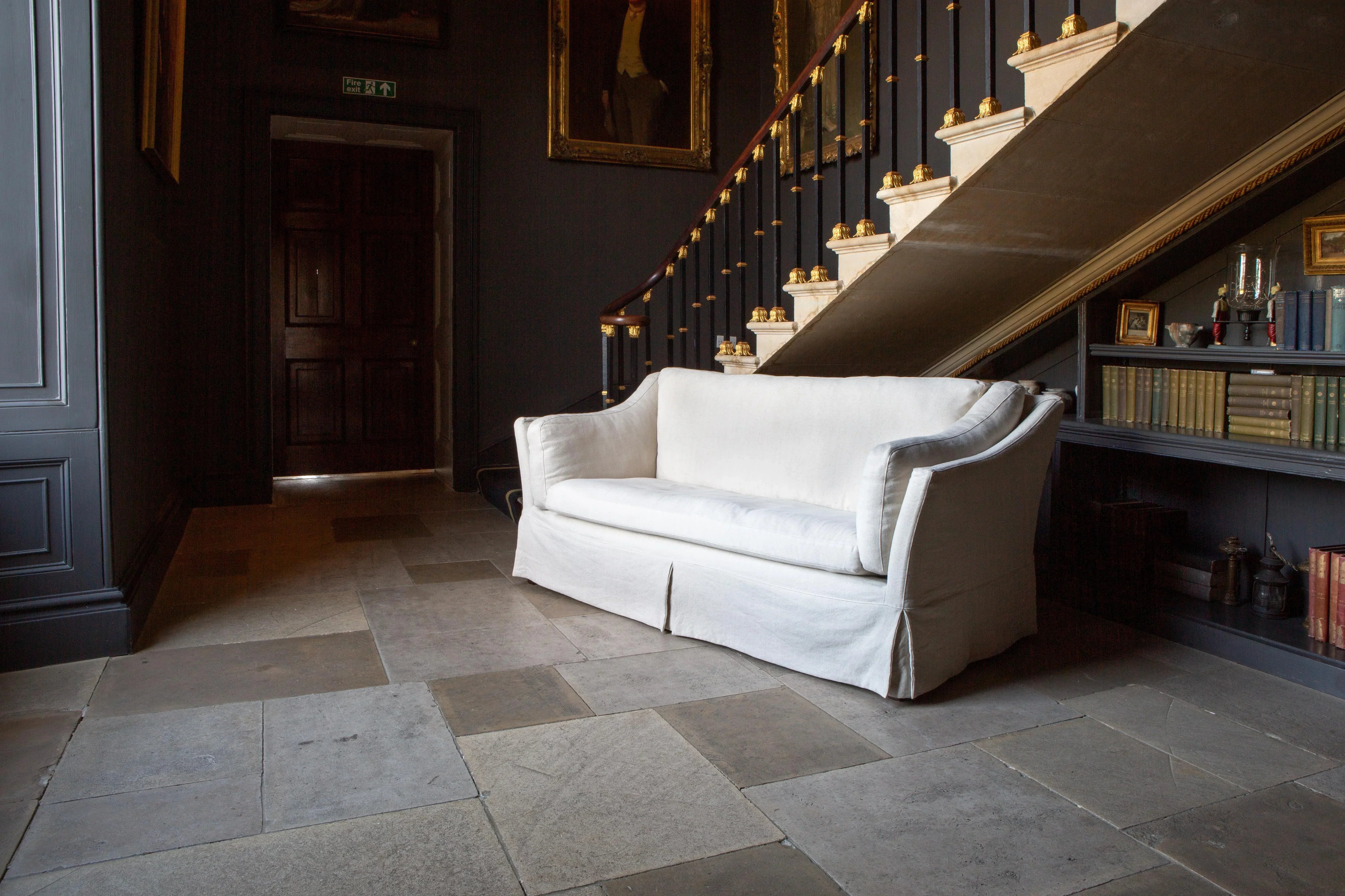 White fabric sofa with skirted base placed in a stone hallway beside a staircase and bookshelf