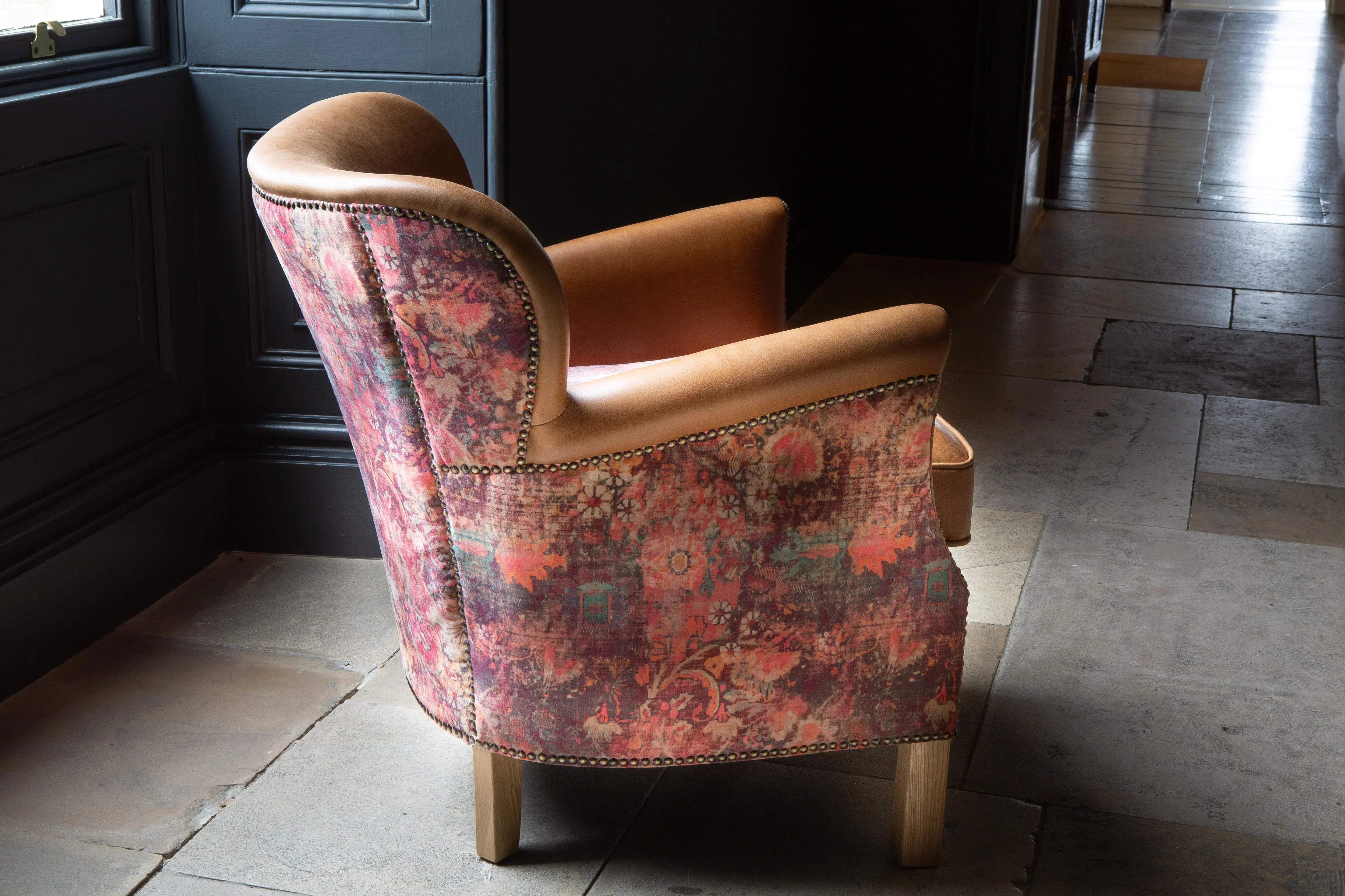 Patterned upholstered armchair with tan leather seat and wooden legs by window on stone floor