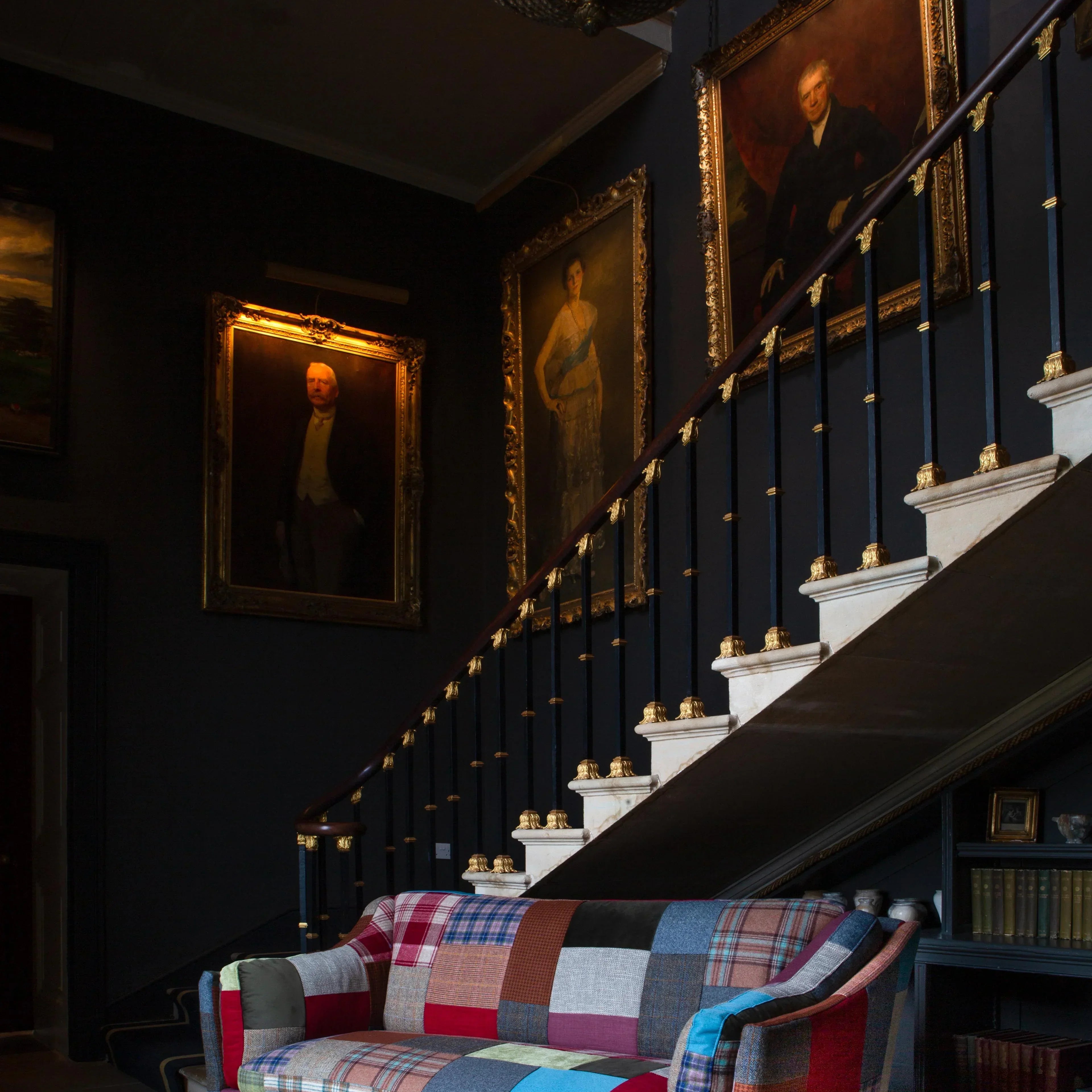 Patchwork fabric sofa in a classic hallway with dark walls, gold-framed portraits, and staircase