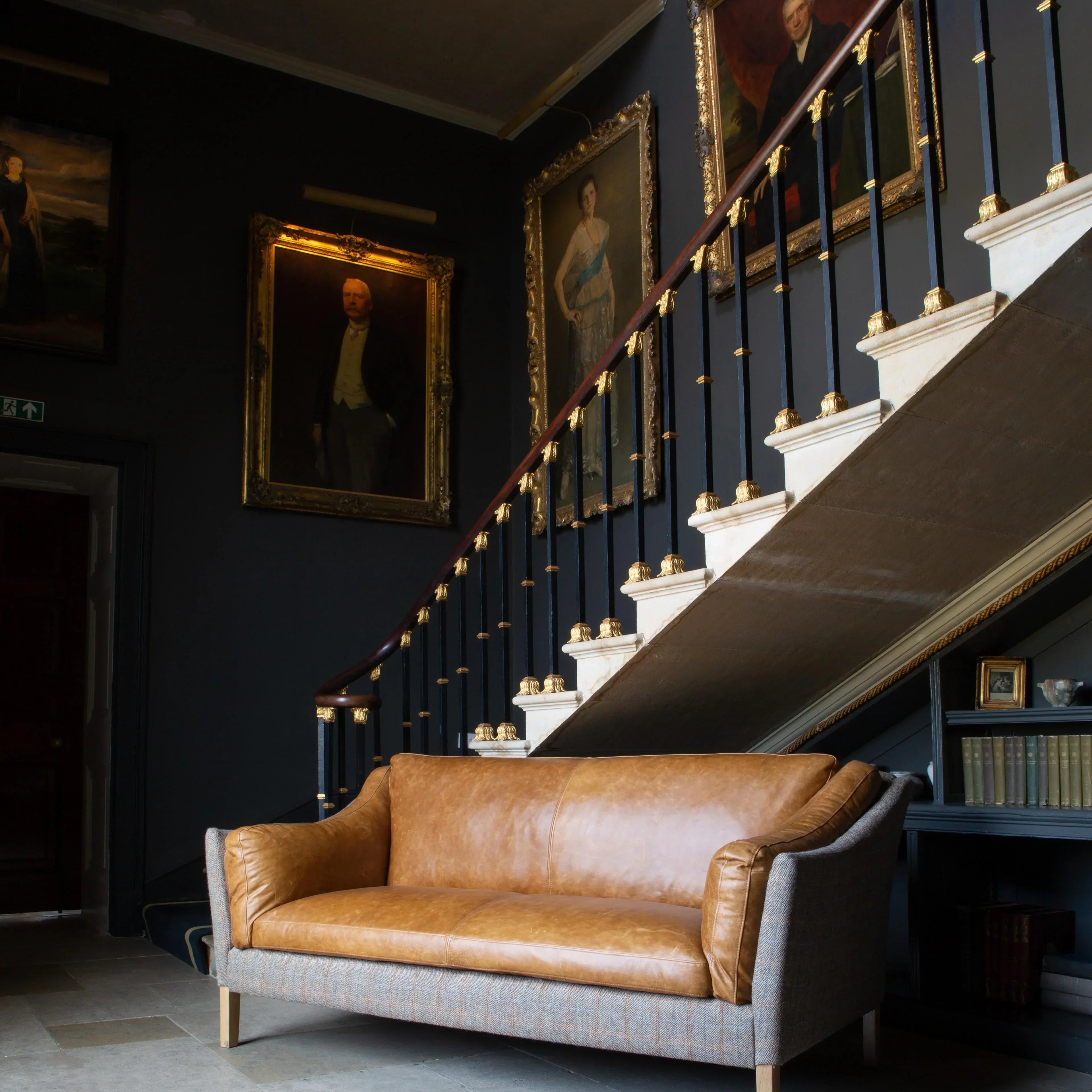 Tan leather and fabric sofa in front of staircase with framed portraits and chandelier