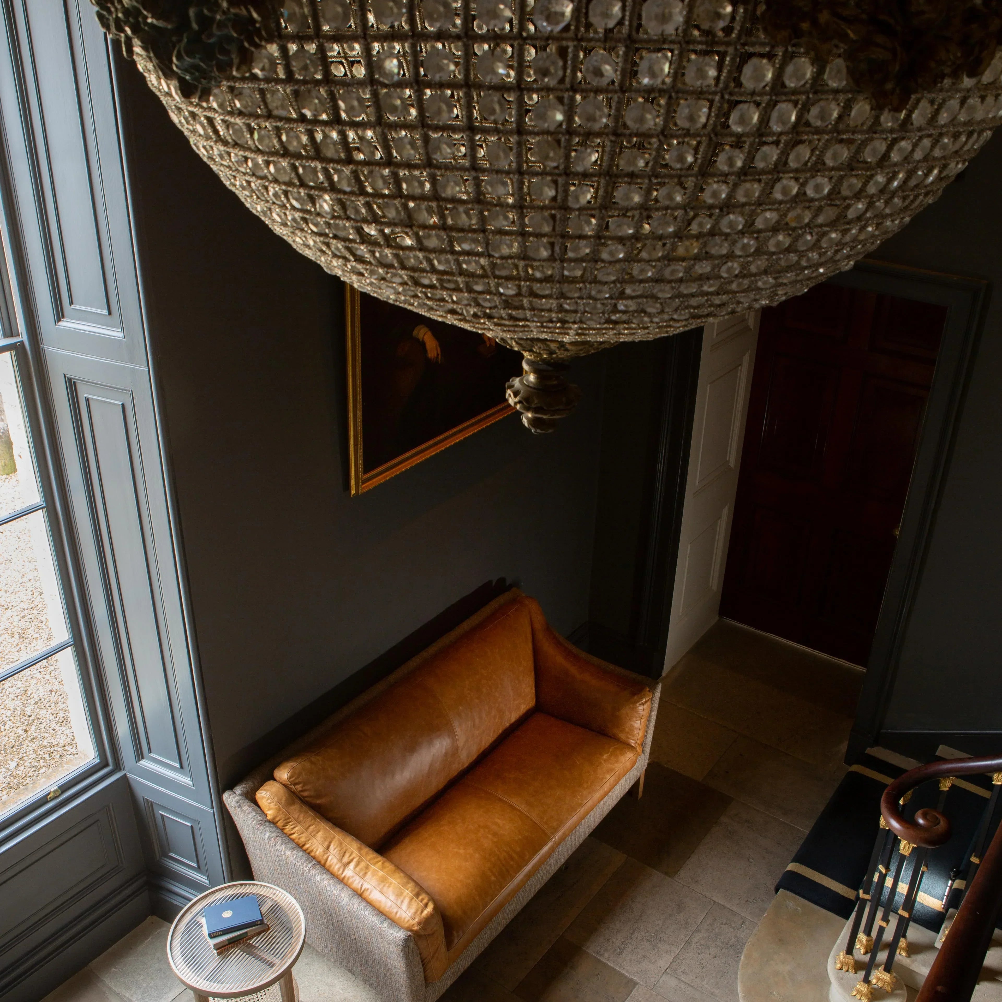 Tan leather sofa with light wood frame in entryway beside round side table and chandelier