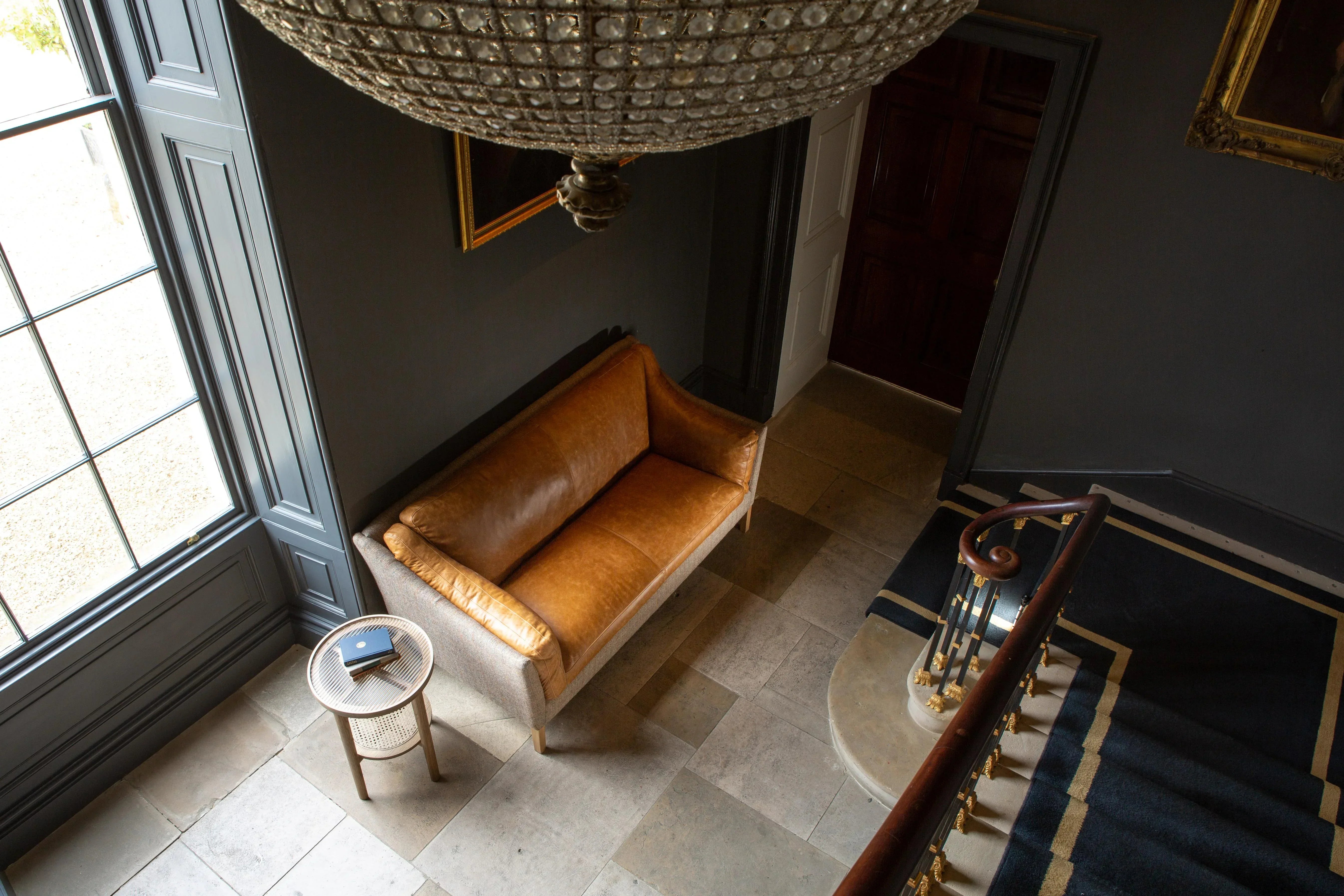 Tan leather sofa in hallway corner beside round cane side table and large window