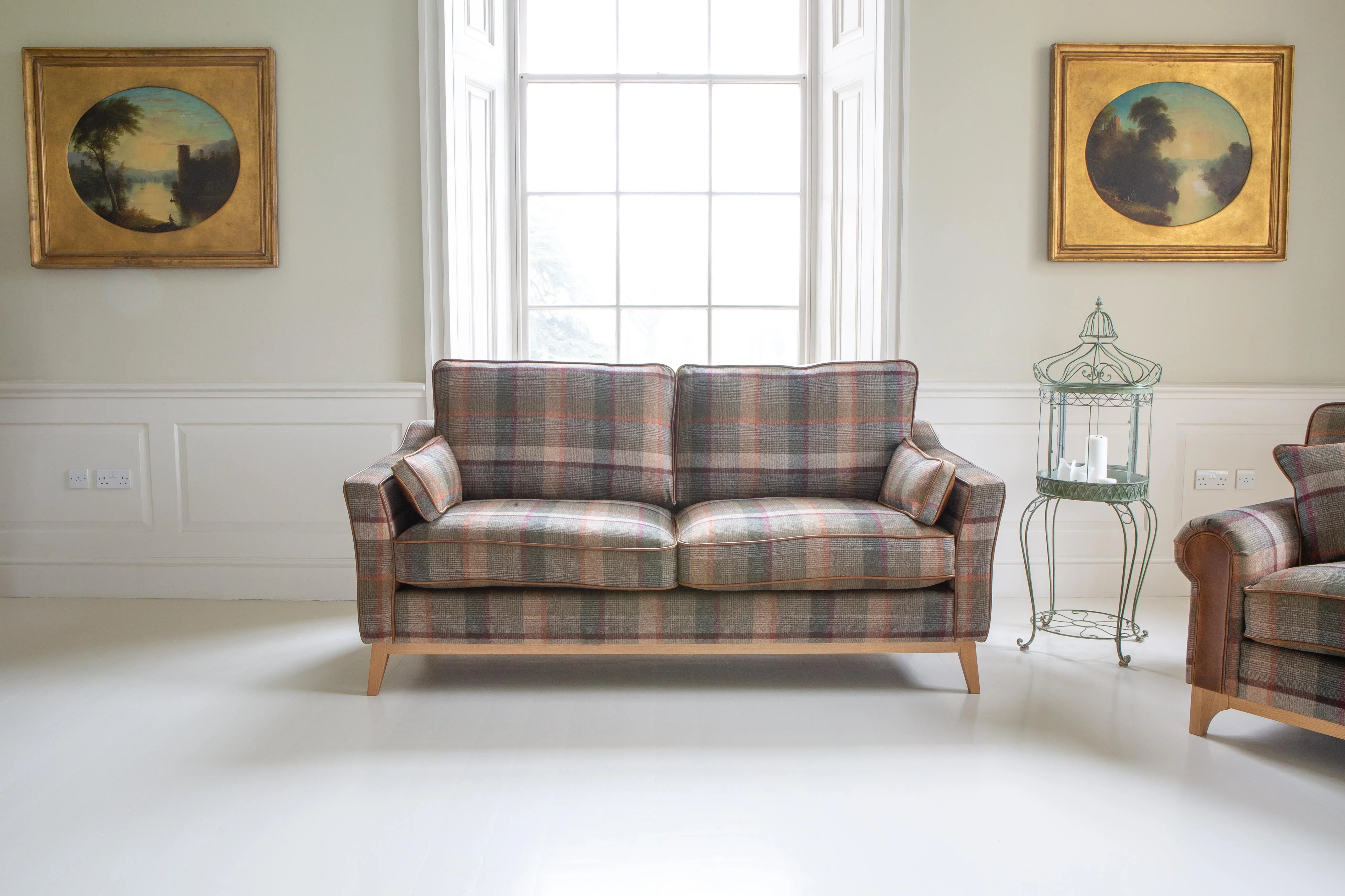 Plaid fabric sofa with wooden legs in bright living room between two framed landscape paintings