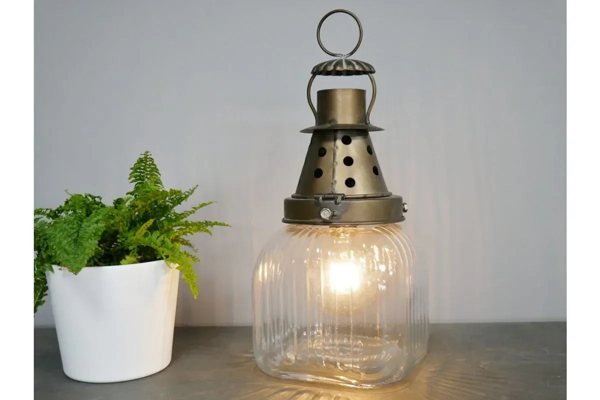 Vintage glass lantern table lamp with metal top beside potted fern on grey surface