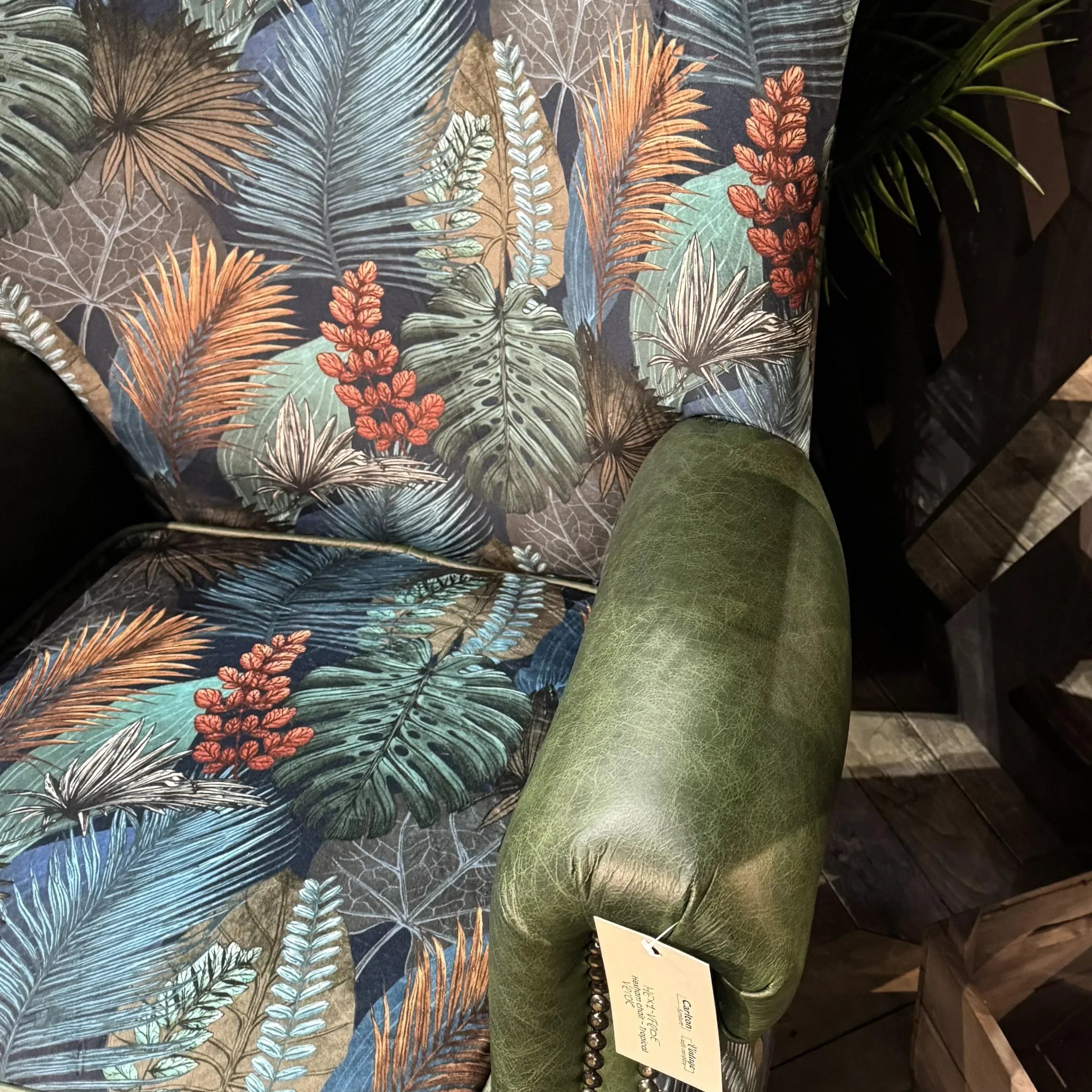 Patterned armchair with tropical leaf upholstery and green leather armrest on wooden floor