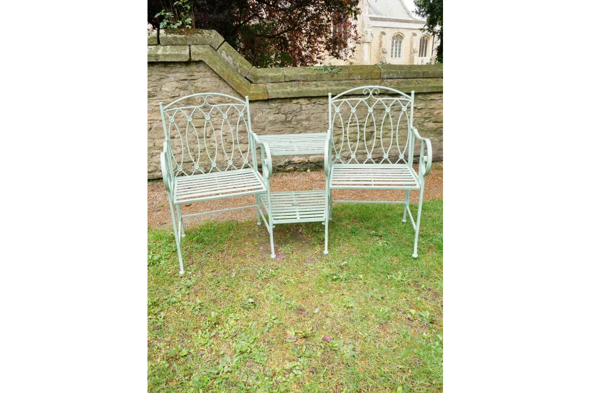 Green metal garden loveseat bench with central table on grass near stone wall