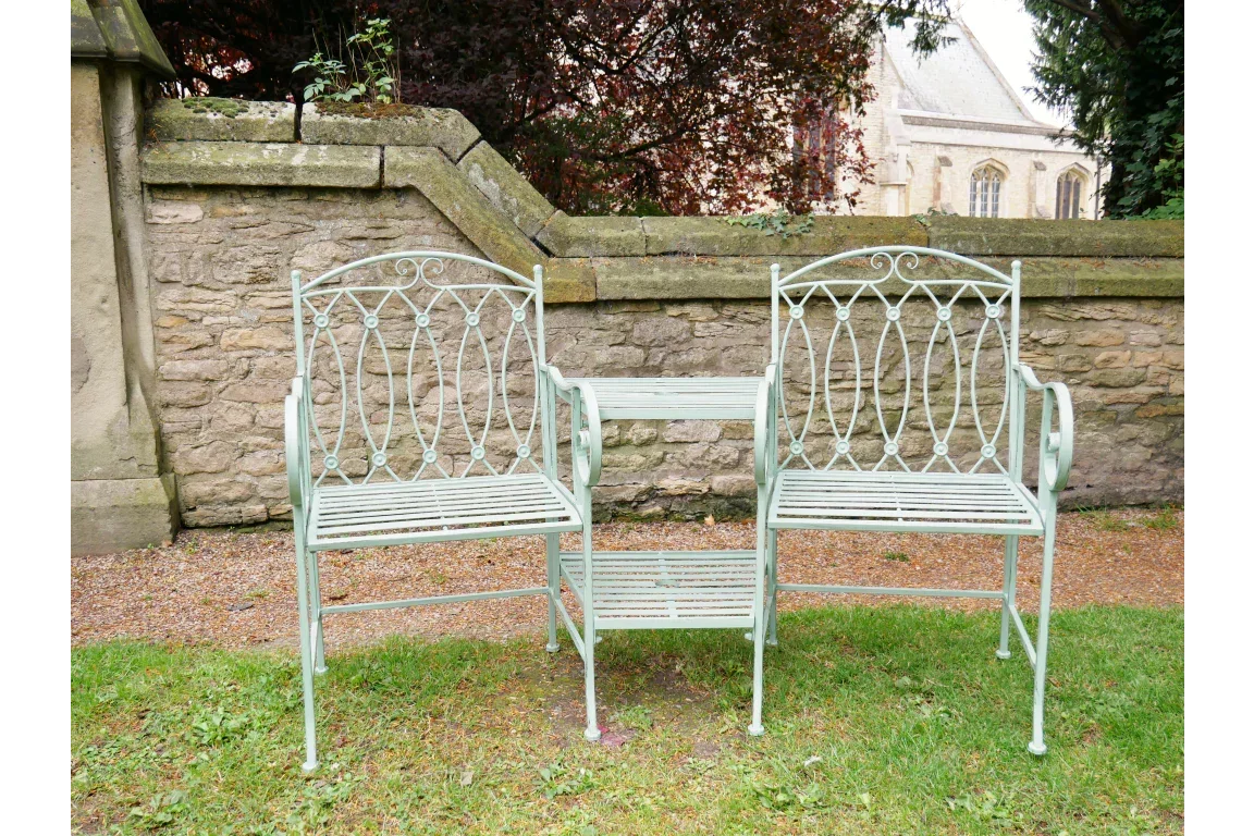 Metal garden love seat with table, powder-coated pale green, set on grass by stone wall